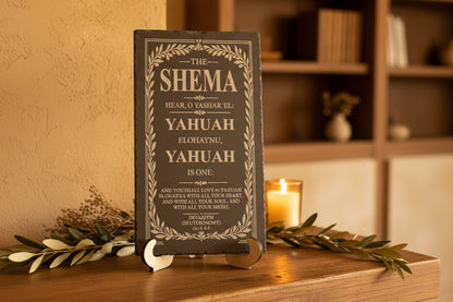 Decorative Shema slate tablet with engraved text on a stand, placed on a wooden surface with a candle and foliage.